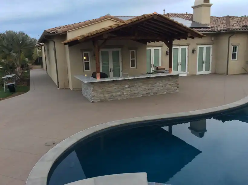 Large concrete pool deck curving around a pool toward an outdoor kitchen bar and pergola