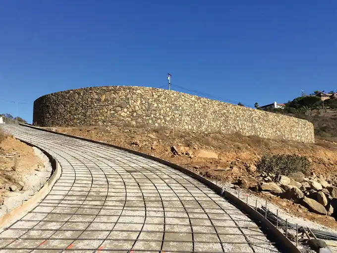 Curving walkway laid out in a tied rebar grid leading to a stone-walled round feature under a clear sky